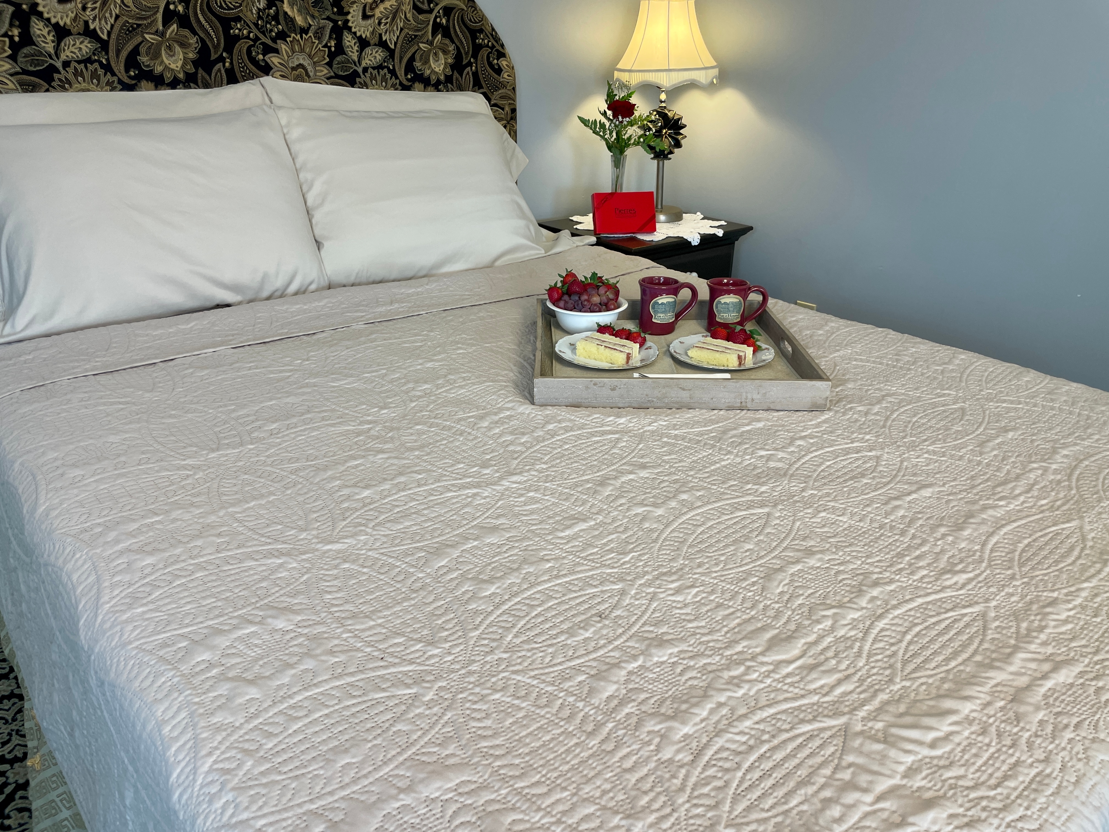 Queen bed with tan coverlet and tray of cakes and coffee on bed with red rose in a vase