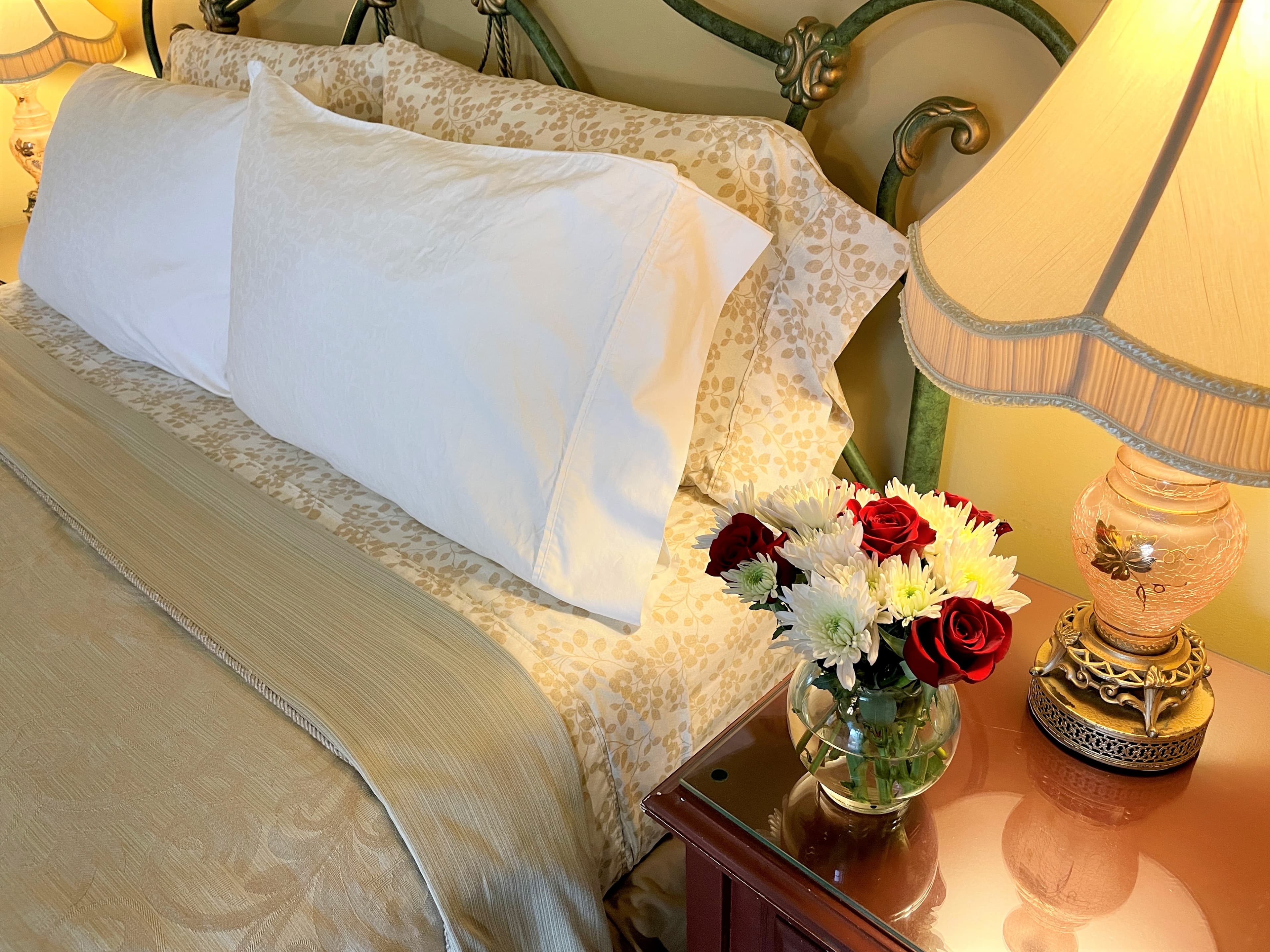 nightstand with vintage pink glass lamp and small vase of fresh roses next to white ironed pillowcases