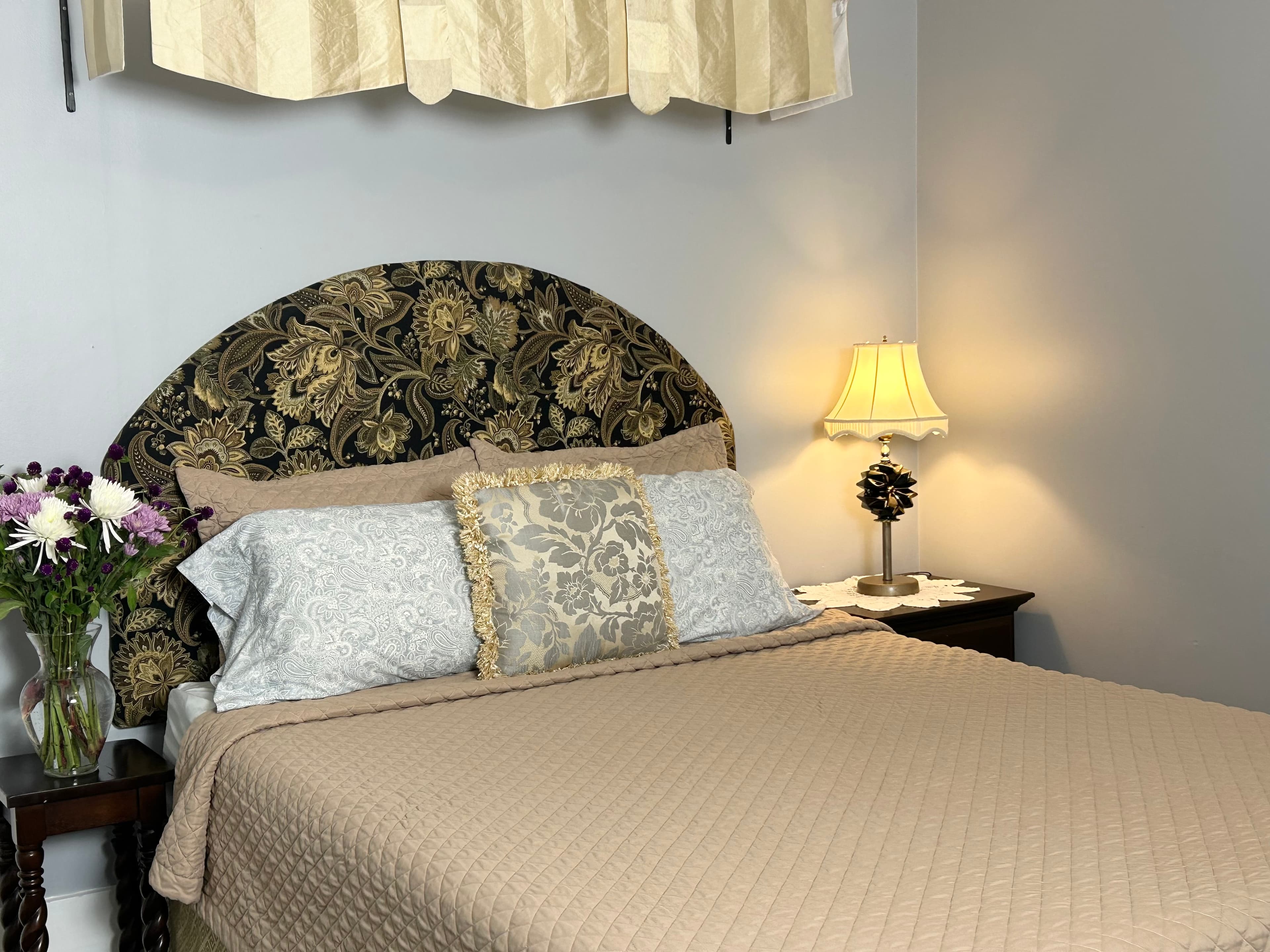 Queen bed with flowers on the nightstand and a lamp with fabric wall hanging