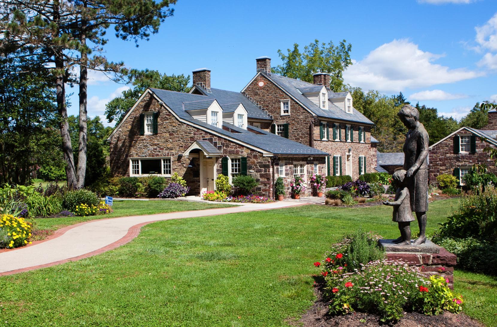 A stone house with a manicured garden and a statue of a woman and child in front. A stone house with a manicured garden and a statue of a woman and child in front.