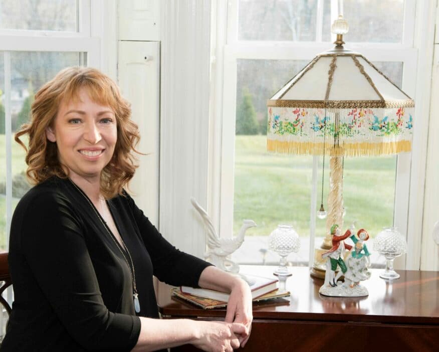 A woman with curly hair smiles while sitting at a table next to a lamp and decorative figurines.
