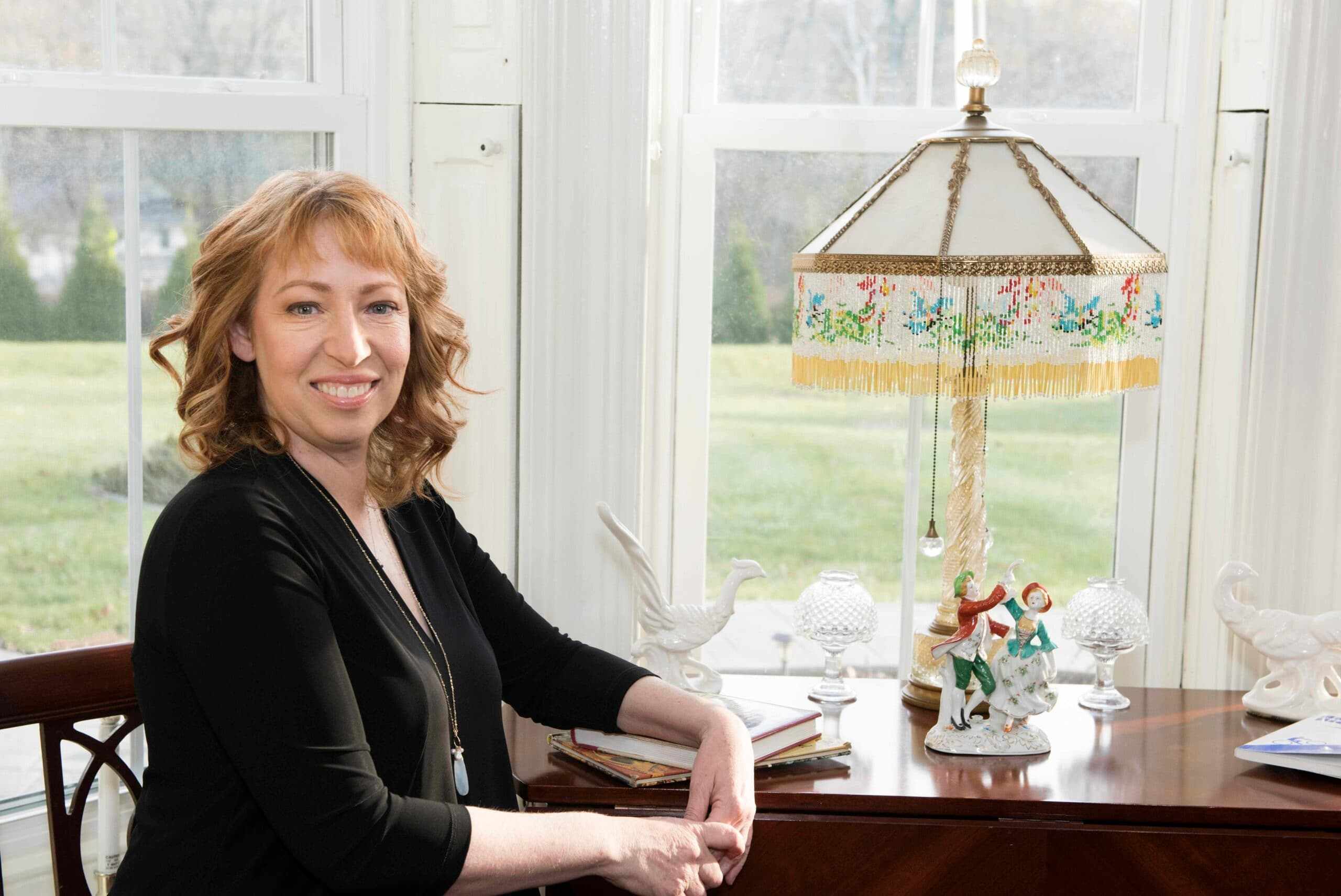 Innkeeper sitting in Parlor next to a vintage lamp and some Cookbooks on a sunny day.